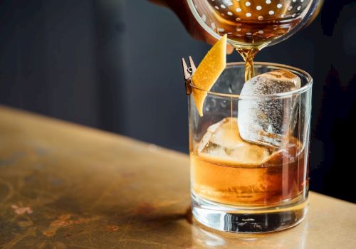 A neat whiskey on the rocks being poured into a glass, with an orange twist garnish and a chilled ice cube holding a glassy silhouette.