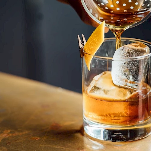 A neat whiskey on the rocks being poured into a glass, with an orange twist garnish and a chilled ice cube holding a glassy silhouette.
