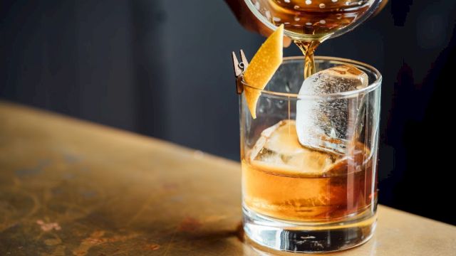 A neat whiskey on the rocks being poured into a glass, with an orange twist garnish and a chilled ice cube holding a glassy silhouette.