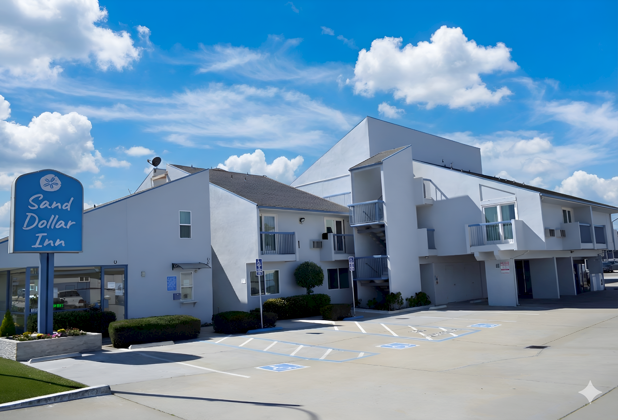 A small roadside motel with a blue sign reading &ldquo;Sand Dollar Inn,&rdquo; white two-story buildings, parking spots, and a bright blue sky with clouds.
