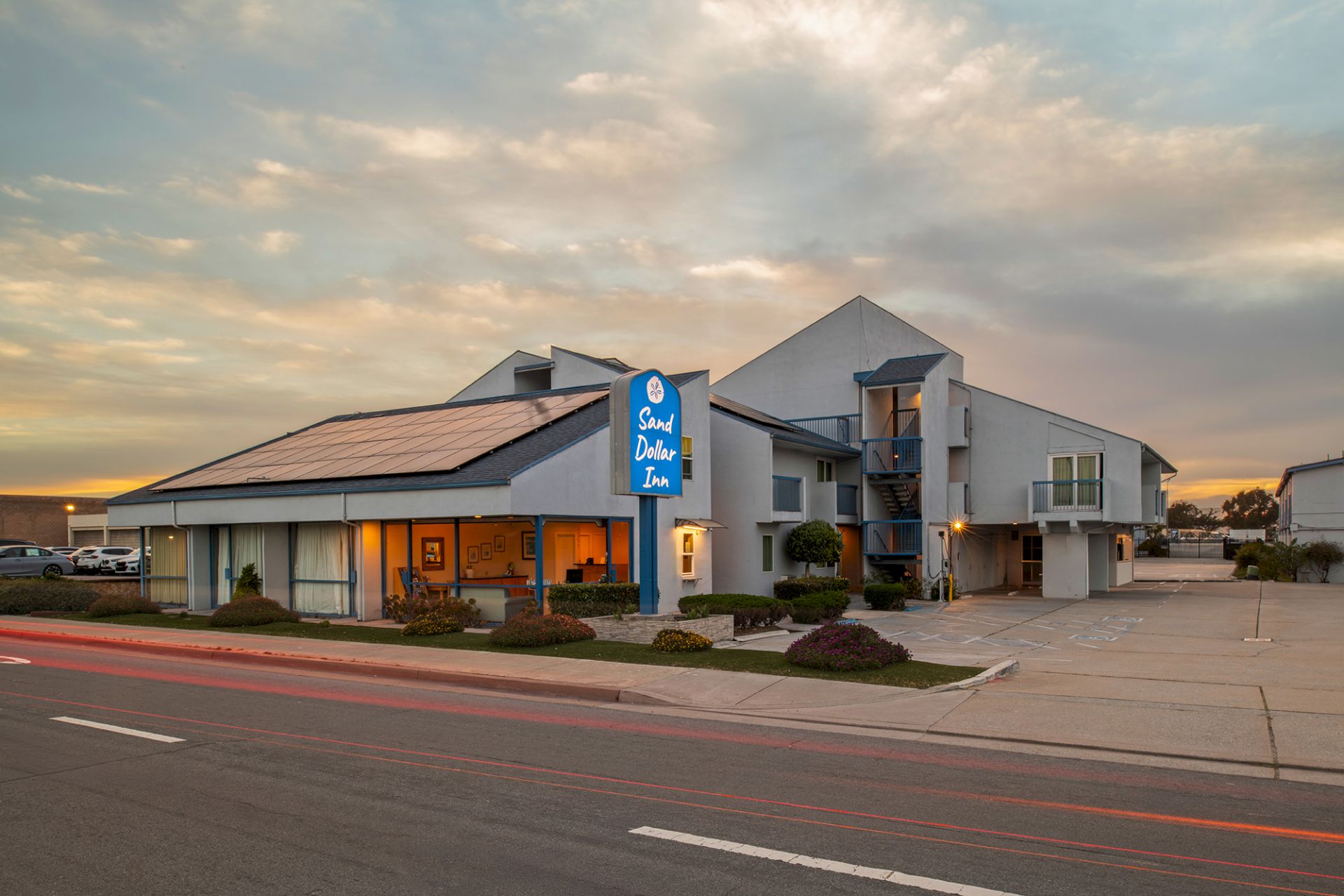 A small motel at sunset with a blue &ldquo;Quality Inn&rdquo; sign, paved parking, and a modern two-story building near a busy street.