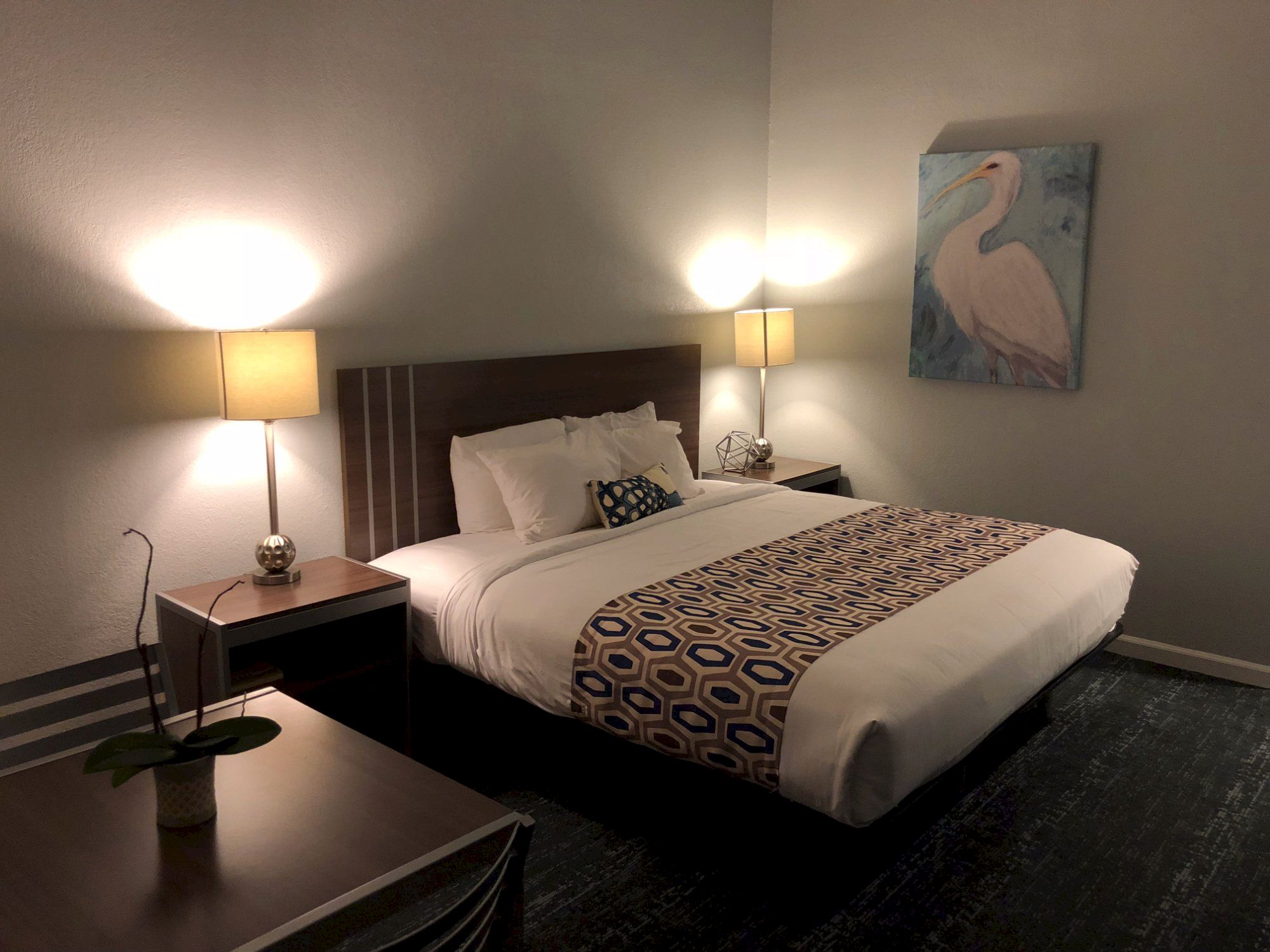 A hotel bedroom with a king bed, two nightstands, lamps, a desk, and a painting of a heron on the wall, cozy and clean.