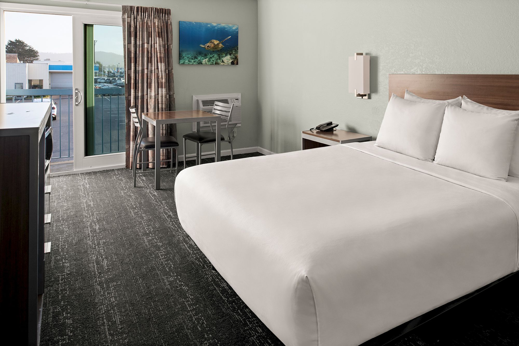 A hotel room with a large bed, a small table and chairs, a balcony door, a dresser, and a wall-mounted artwork.