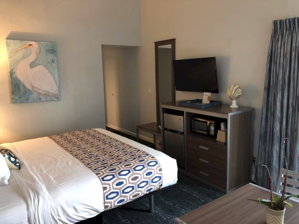 A hotel-style bedroom with a queen bed, geometric bedspread, wall art of a swan, dresser with flat-screen, mirror, and blue-gray decor.