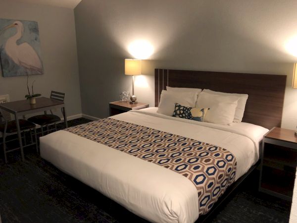 A hotel room with a king bed, two nightstands, lamps, a patterned bed runner, and a small desk with a chair near a pelican artwork.