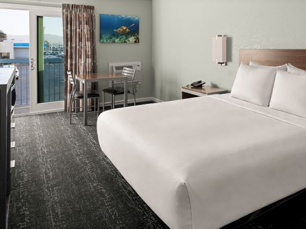A clean hotel room with a large bed, a small table and chairs near a sliding door to a balcony, and an ocean view painting on the wall.