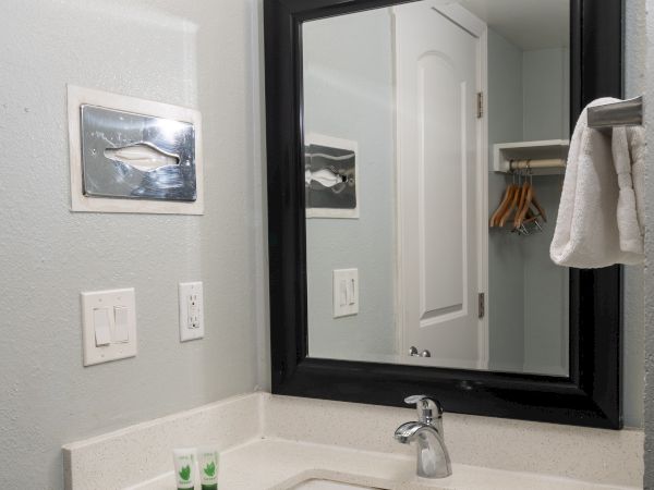 A small bathroom vanity with a white countertop, rectangular sink, dark-framed mirror, three-light fixture, and a towel on a hook in the open closet reflected in the mirror.
