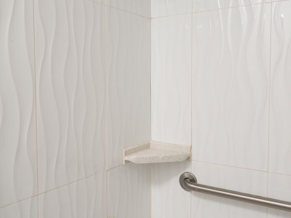 A white tiled shower with a chrome showerhead, a soap shelf, a bath spout, and a silver grab bar along the wall.