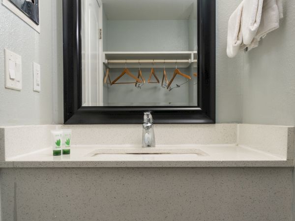 A small bathroom vanity with a single sink, mirror, lights, and a glimpse of a closet through the reflection, clean and minimal in style.