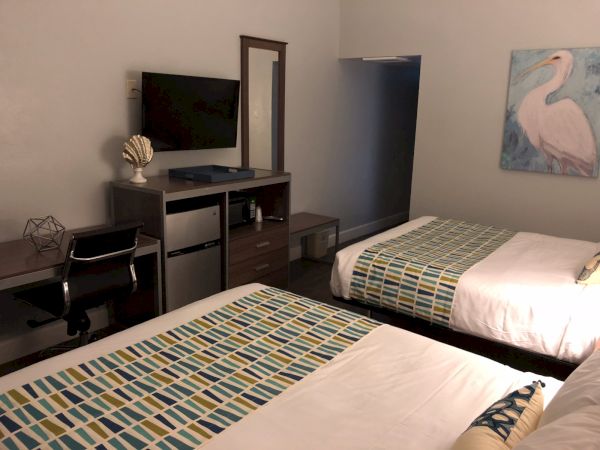 Two double beds with colorful geometric runners, a desk with a chair, a wall-mounted TV, a small dresser, and a painting of a flamingo in a simple hotel room.