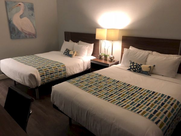 Two twin beds in a hotel room with a nightstand, lamp, decorative pillows, and a stitched geometric runner; a wall art of a bird hangs nearby.