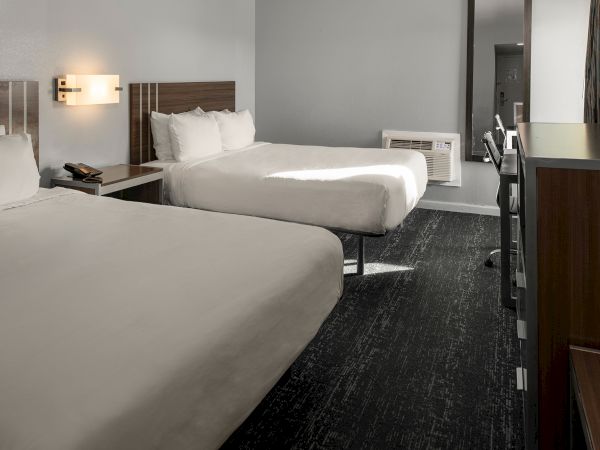 Two hotel beds, nightstands with lamps, a desk and chair, a mirror, and a dark carpet in a modern guest room, with neutral colors.