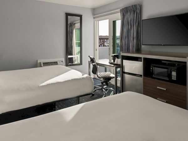 Two neatly made hotel beds, a desk with a chair, a wall-mounted TV, and a cabinet with a microwave; balcony doors let in light. (ends with a period)