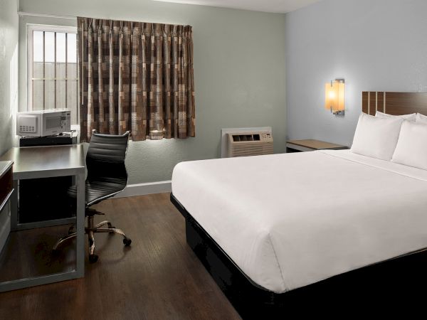 A clean hotel room with a king bed, white linens, a desk and chair, a window with patterned curtains, and a small TV area, all neatly arranged.