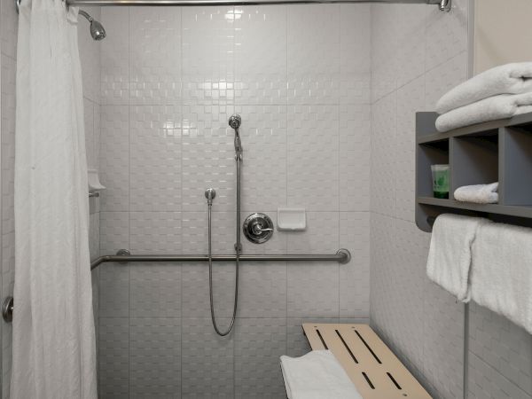A accessible shower area with a handheld shower, a sliding white curtain, tiled walls, a wooden bench, and a shelf with folded towels.