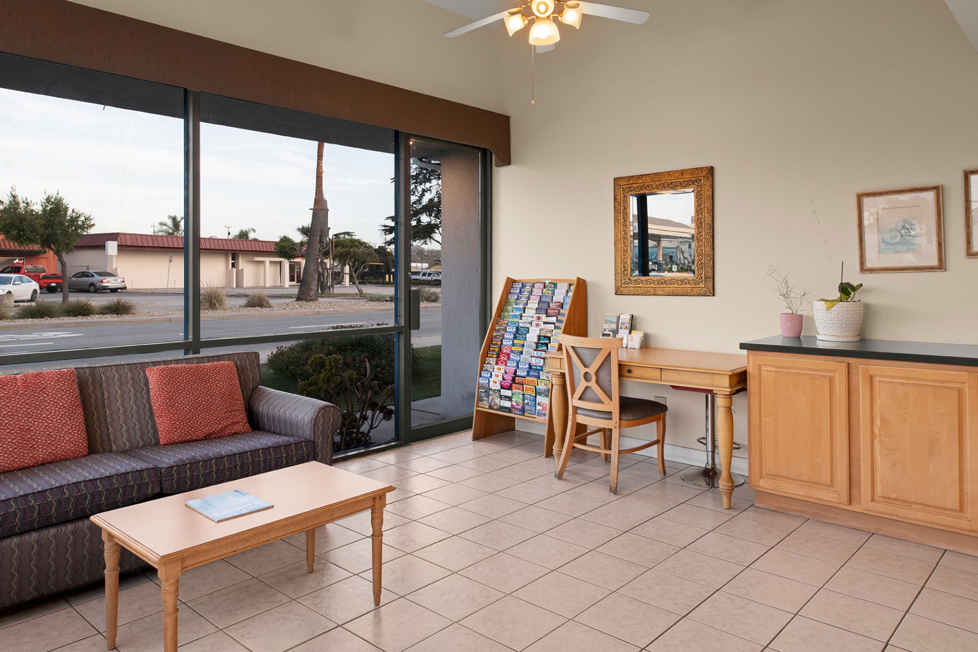 A modern, sunlit lobby or lounge with large windows, a striped sofa, a coffee table, a small desk and chair, cabinetry, and a tiled floor, ending with a ceiling fan.