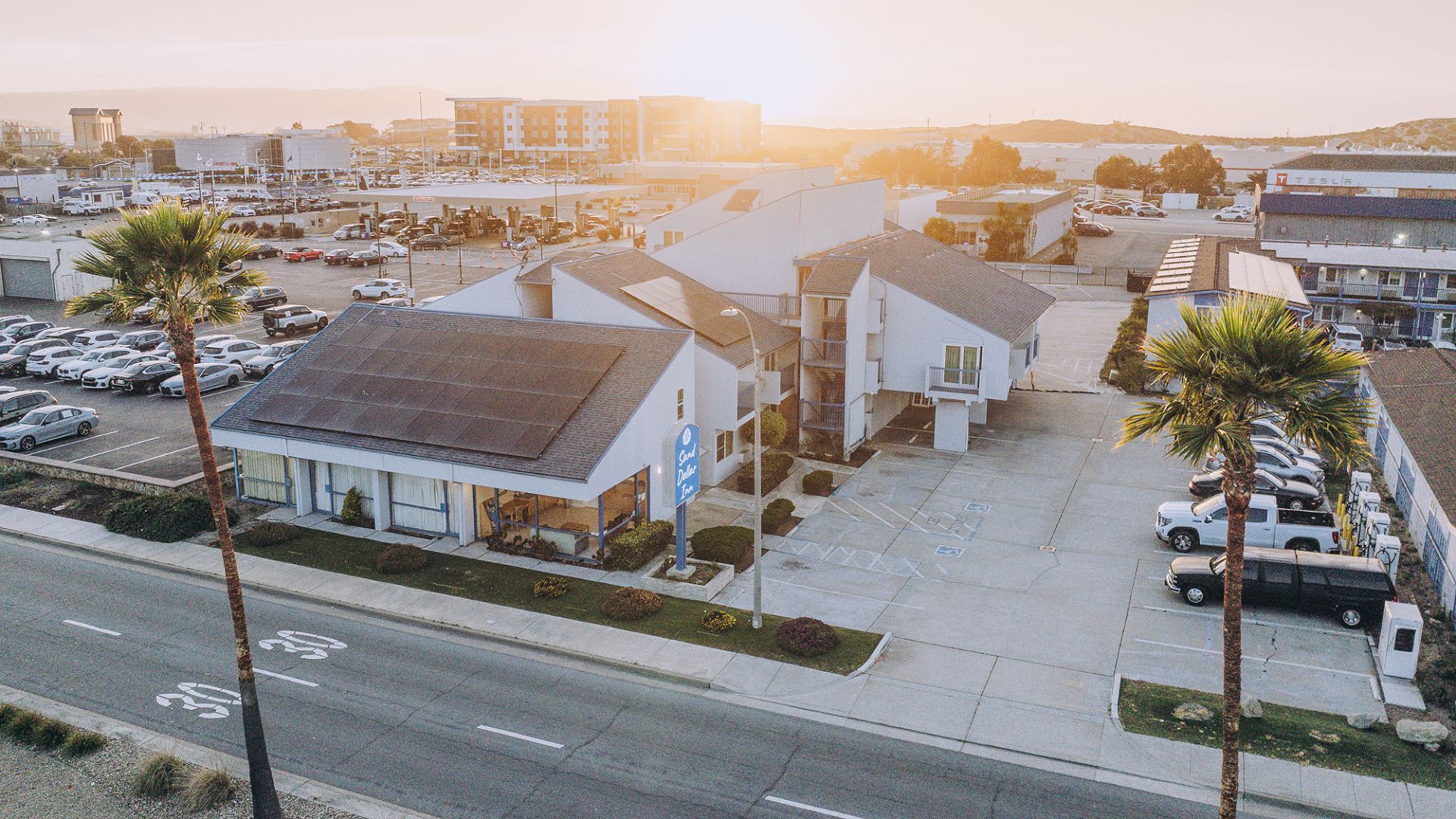 Aerial view of a small coastal town with low-rise buildings, palm trees, a sunny sky, and a parking lot near a road.