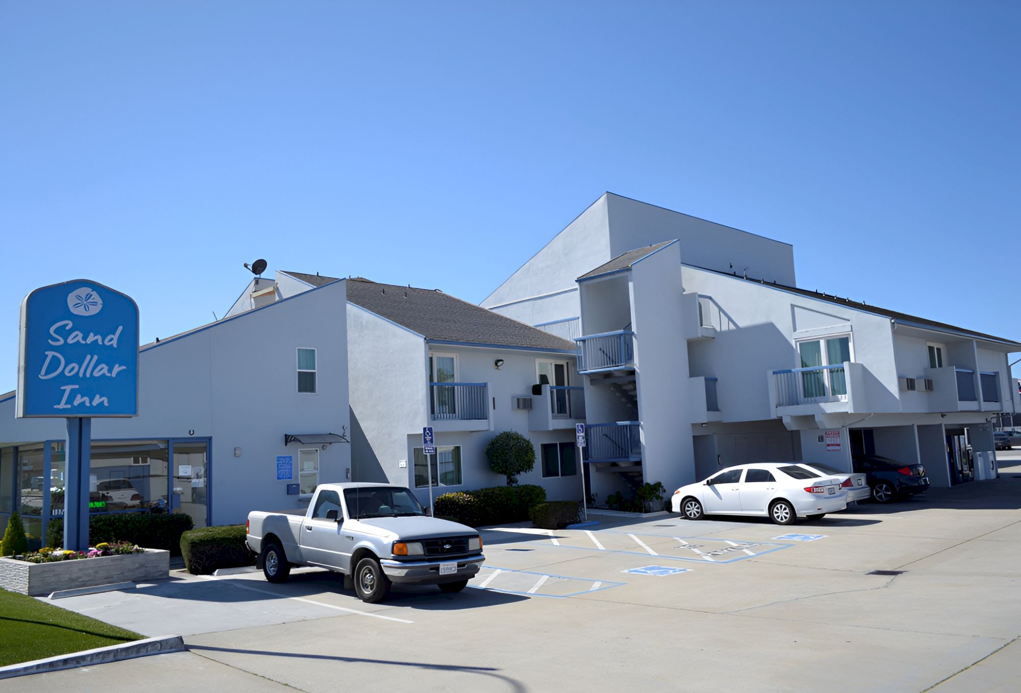 A roadside motel with a blue sign reading &ldquo;Sand Dollar Inn,&rdquo; white two-story buildings, a few parked cars, and a clear blue sky.