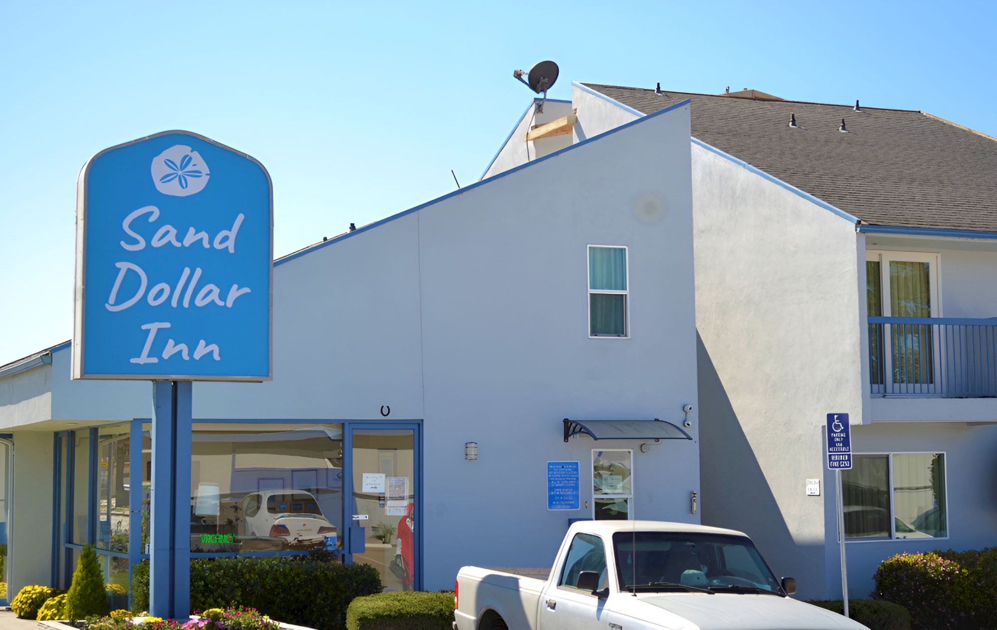 A Sand Dollar Inn building with a blue sign, a white pickup parked outside, and a two-story motel-style exterior under a clear sky.