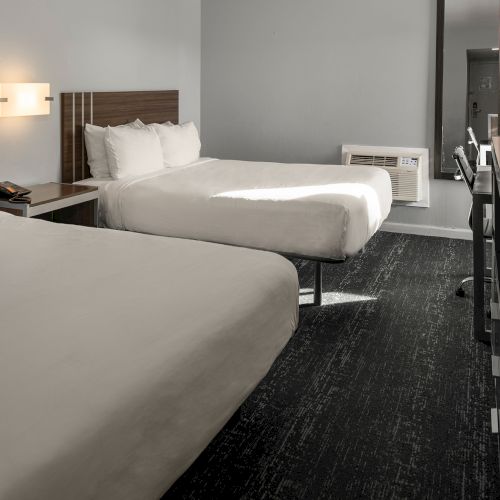 A hotel room with two neatly made beds, nightstand with a lamp, a desk and chair, a mirror, and a carpeted floor.