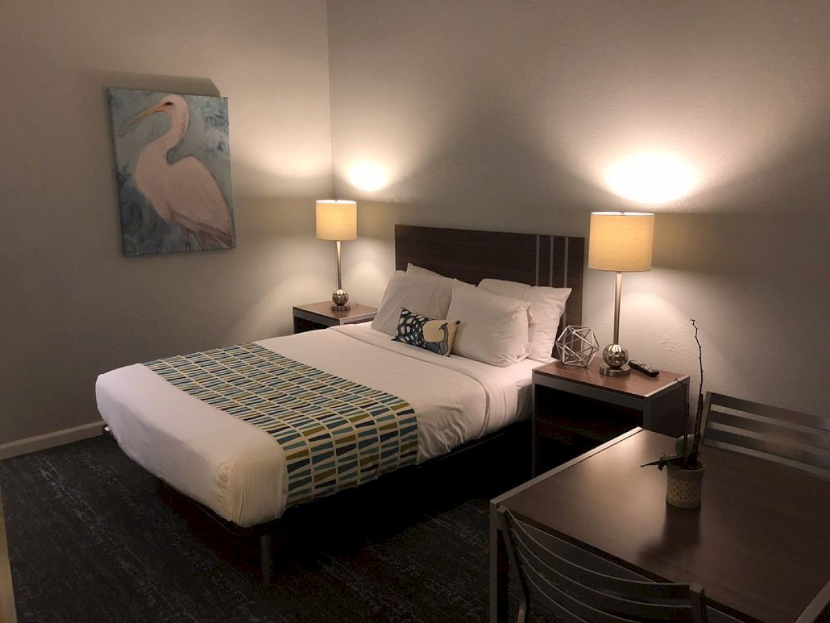 A cozy hotel room with a king bed, two nightstands, lamps, a framed swan artwork, and a small desk with a chair. Ends here.