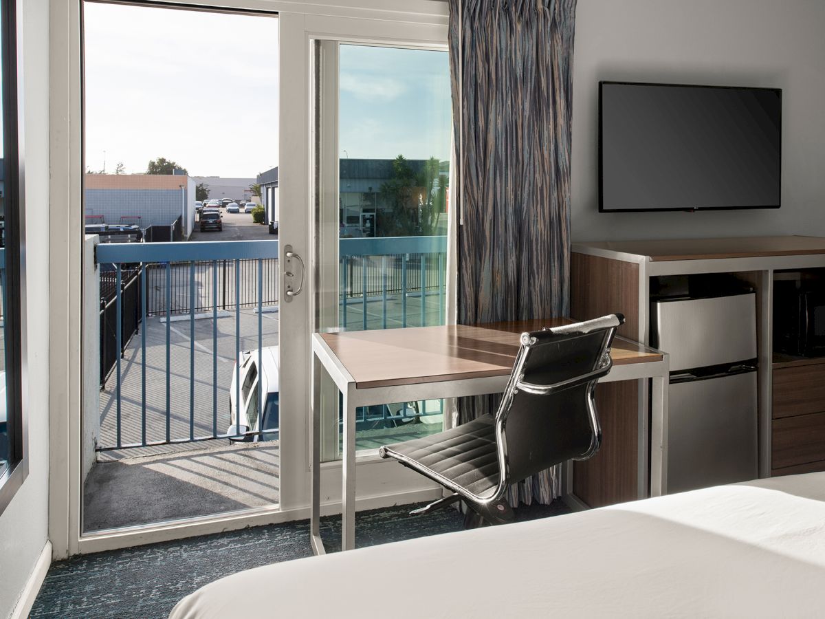 A bright hotel room with a desk and chair, large balcony doors open to a view, a wall-mounted TV, and a mini fridge area. End.