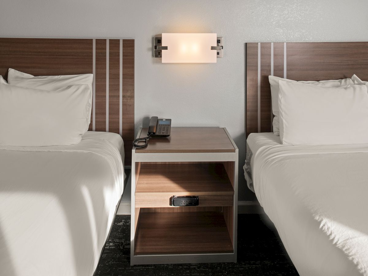 Two neatly made twin beds with a shared nightstand and a wall-mounted light between them, in a simple hotel room.