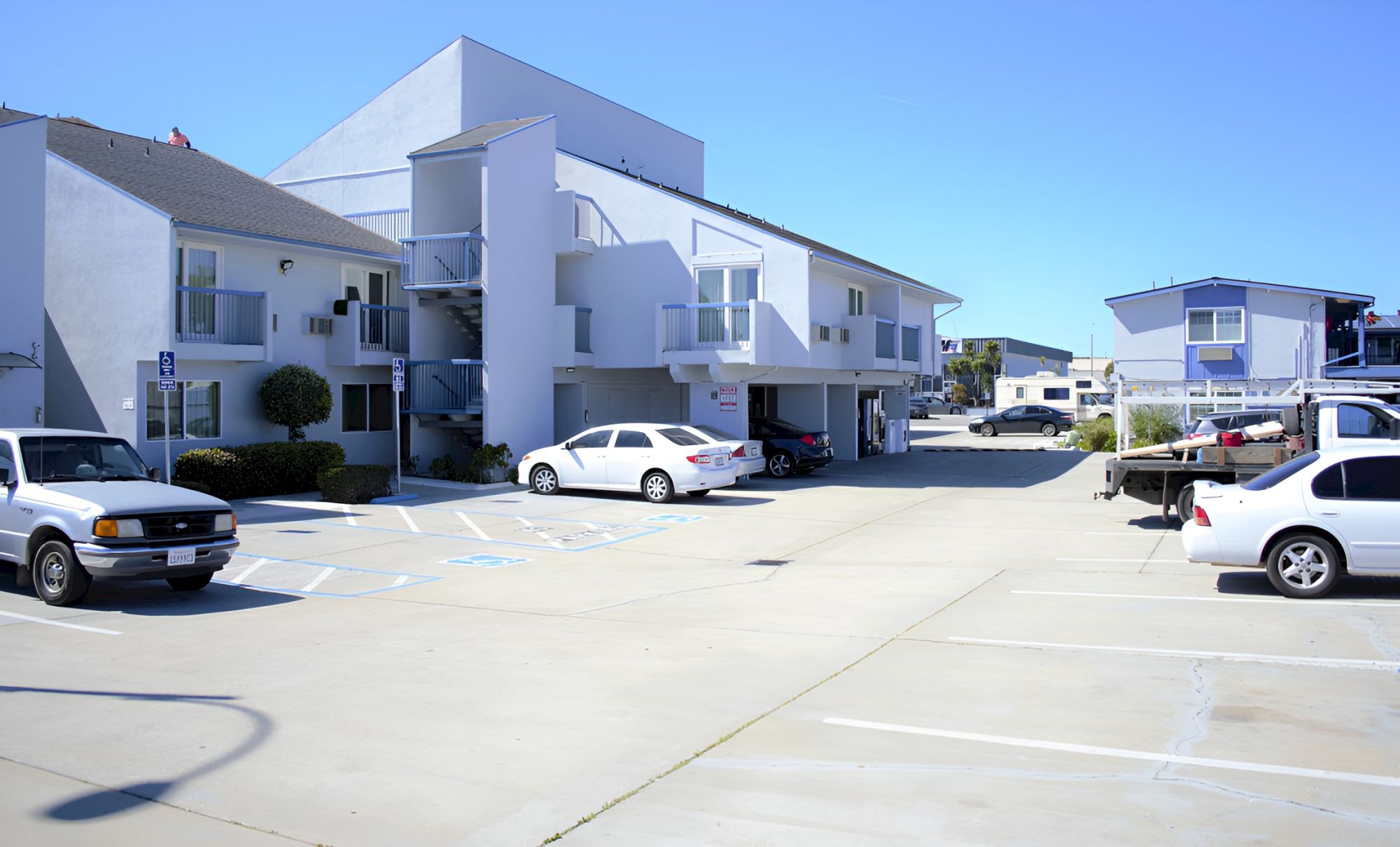 A cluster of light-blue multi-story apartment buildings surrounds a wide, sunlit parking lot with several cars and clear blue skies overhead.