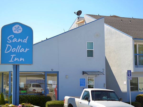 A Sand Dollar Inn with a blue sign, a white two-story building, and a white pickup truck parked out front, in bright daytime.