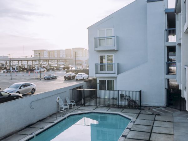 A small rectangular pool on a tiled patio beside a light blue apartment building, with a city street and cars in the background, under a bright sky.