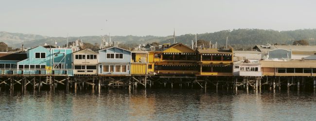 Colorful row of houseboats lined along the water on stilts, pastel blues, greens, yellows, and browns facing a calm harbor.