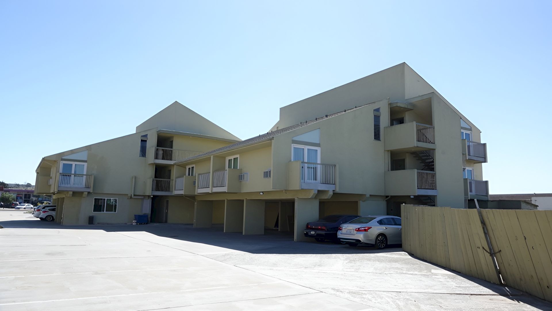 A pastel yellow motel-like building with balconies, a covered walkway, and parked cars in a sunny, open lot.