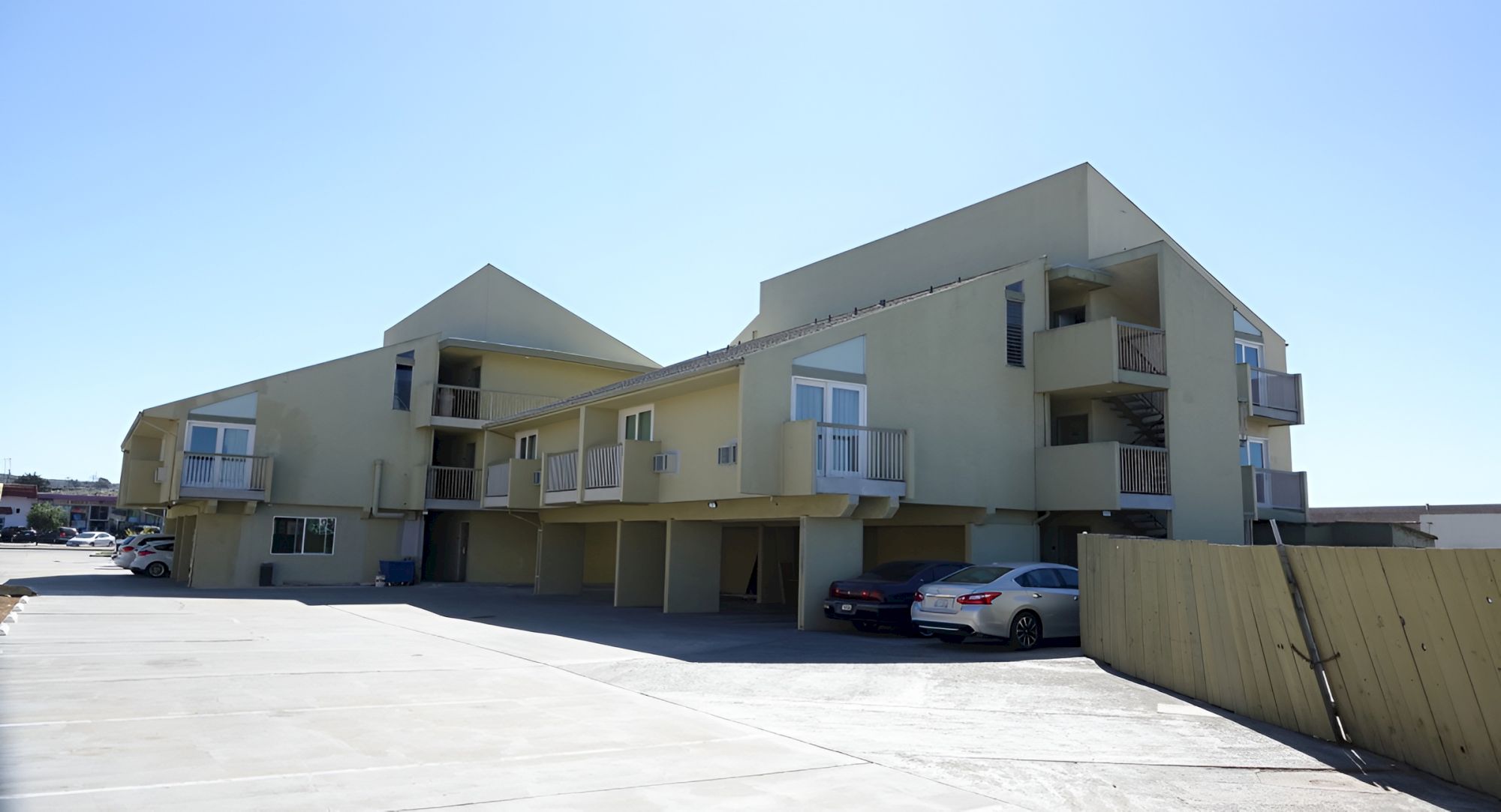 A pastel yellow motel-like building with balconies, a covered walkway, and parked cars in a sunny, open lot.