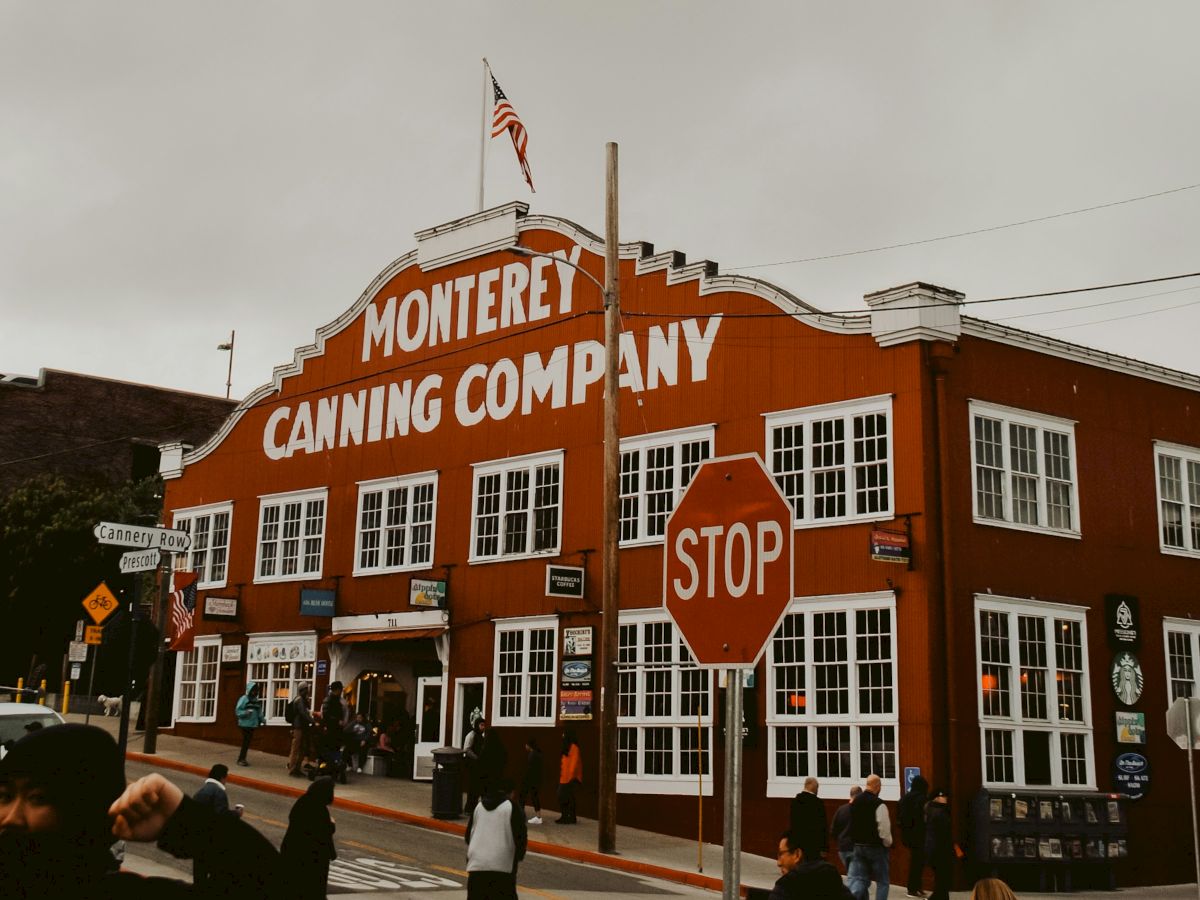 A red brick building labeled &ldquo;Monterey Canning Company&rdquo; with a stop sign, people outside, and power lines overhead at a street corner.