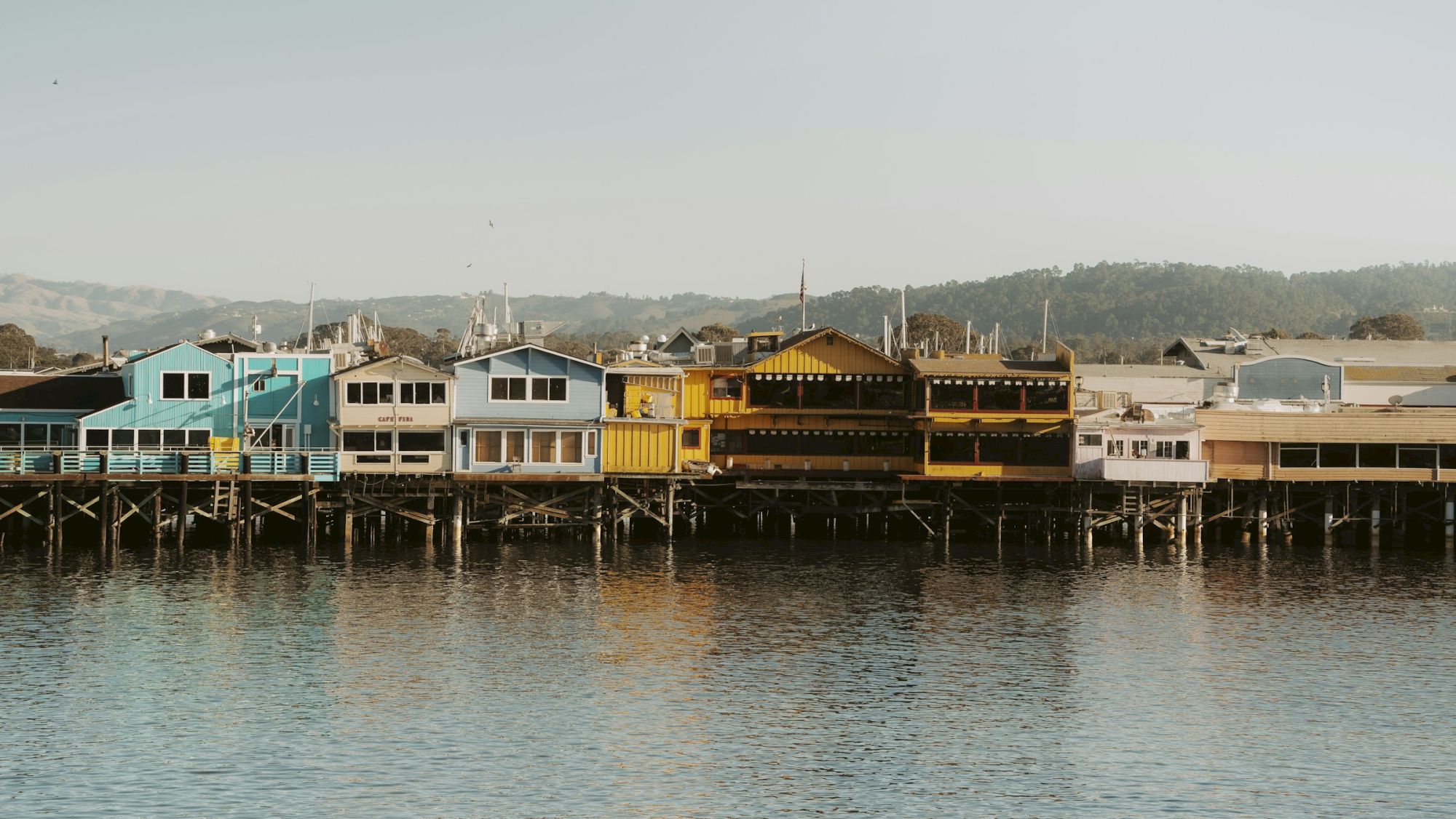 Colorful seaside houses on stilts over calm water, connected along a wooden deck, with hills in the distance.
