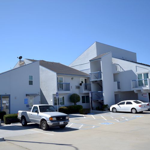 A small auto shop or dealership with a light blue building, a white pickup truck, and a trailer in the lot, sunny day.