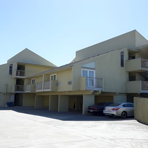 A row of cream-colored townhomes with gabled roofs and small front porches on a sunny day, parked cars lining the street, and a clear blue sky.