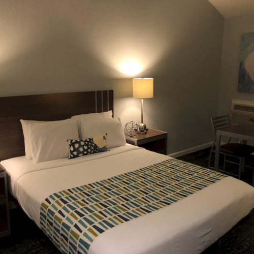 A hotel room with a king bed, two nightstands, lamps, a patterned bed runner, a small desk and chair, and a pelican painting on the wall.