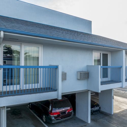 An exterior view of a two-story apartment building with blue railings and covered parking beneath, showing balconies and parked cars.