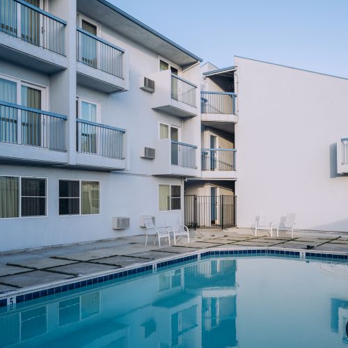 A hotel-style courtyard with a rectangular pool, white building walls, balconies overlooking the pool, and sunlit outdoor seating by the water.