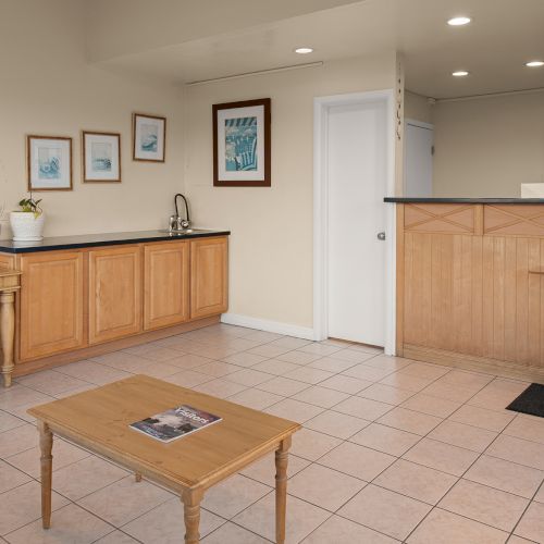 A small, tidy lobby with tiled floor, wooden reception counter, a few cabinets, a side desk, wall art, and a coffee-table in front, bright and welcoming.