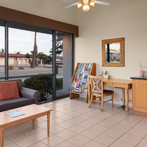 A bright, cozy lobby or common area with a sofa, coffee table, tiled floor, large window, and a small workstation with a chair and cabinets.