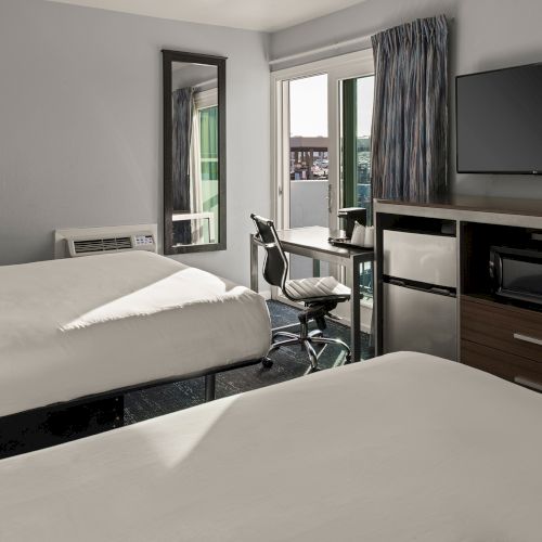 A hotel room with two beds, a desk and chair, a wall-mounted TV, a microwave and mini-fridge unit, and a sliding glass door to a balcony.