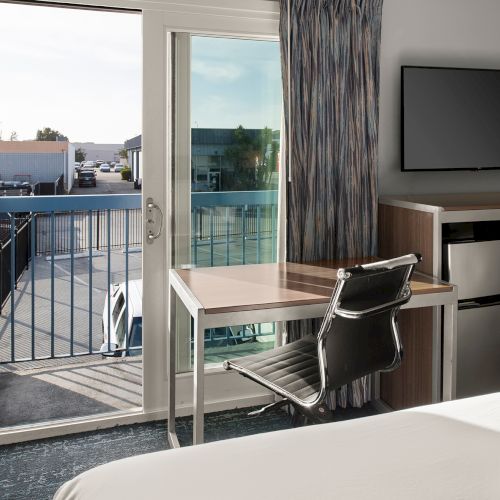 A modern hotel room with a desk by the balcony doors, a chair, a flat-screen TV on a low cabinet, and a small fridge area; balcony views outside.