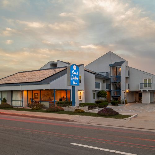 A street view of a modern Sonic-style hotel/inn with a blue sign, glass windows, a parking lot, and a wide street at sunset.