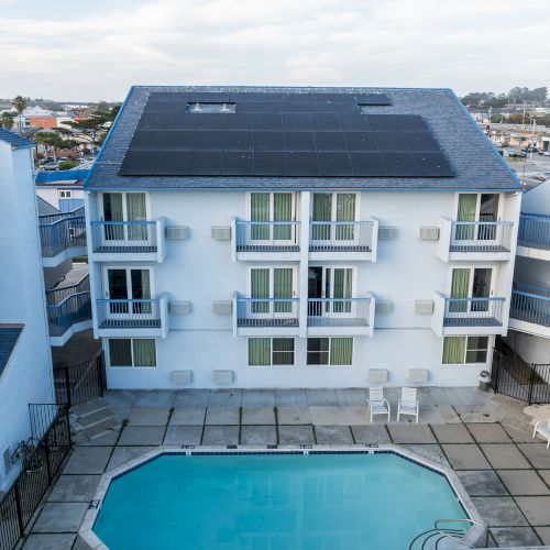 Blue seaside apartment building with a rectangular pool courtyard, balconies facing inward, and clear sky above.