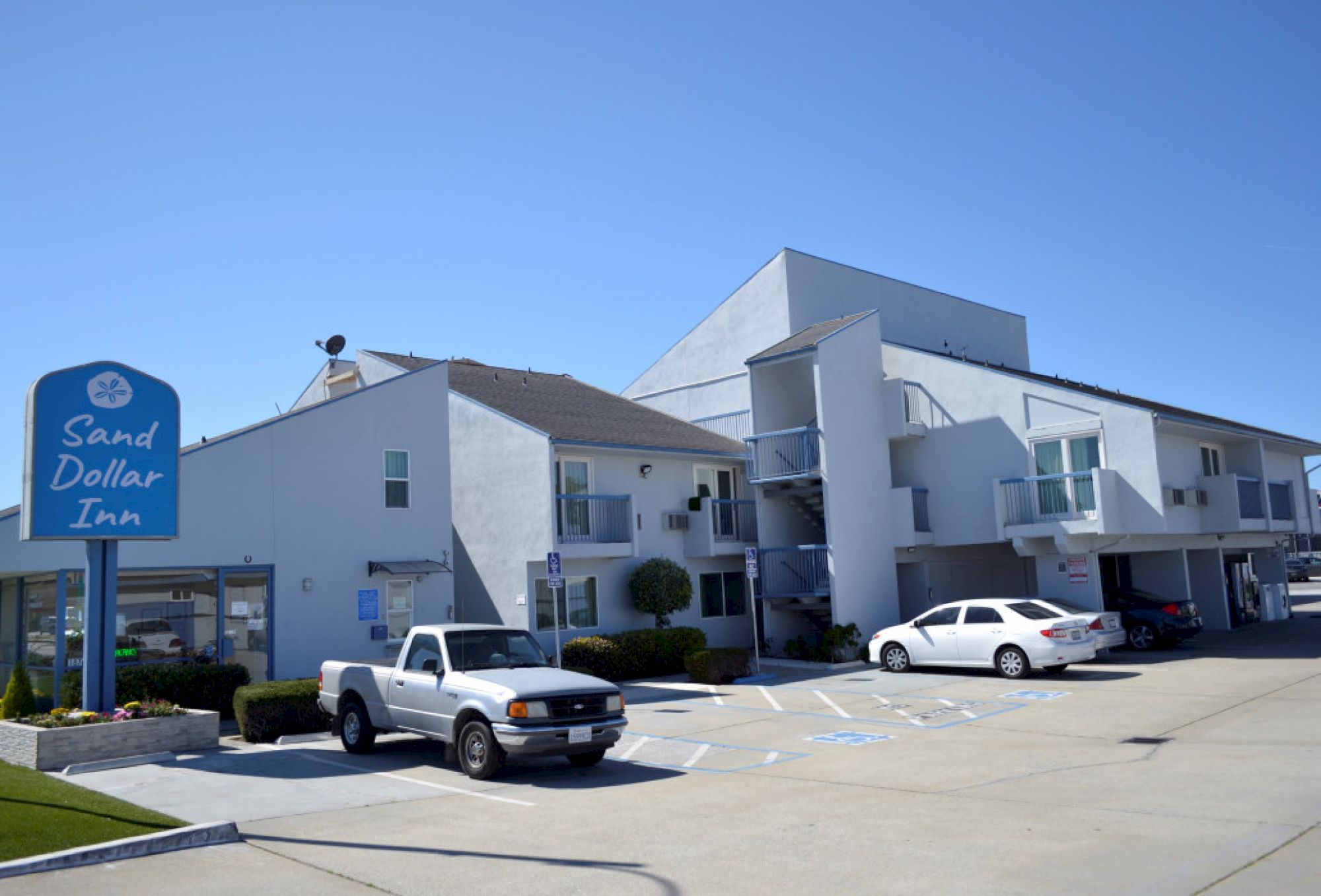 A Budget motel with a blue sign reading &ldquo;Sand Dollar Inn,&rdquo; white units, parked cars, and a bright blue sky ending with a cheerful vibe.