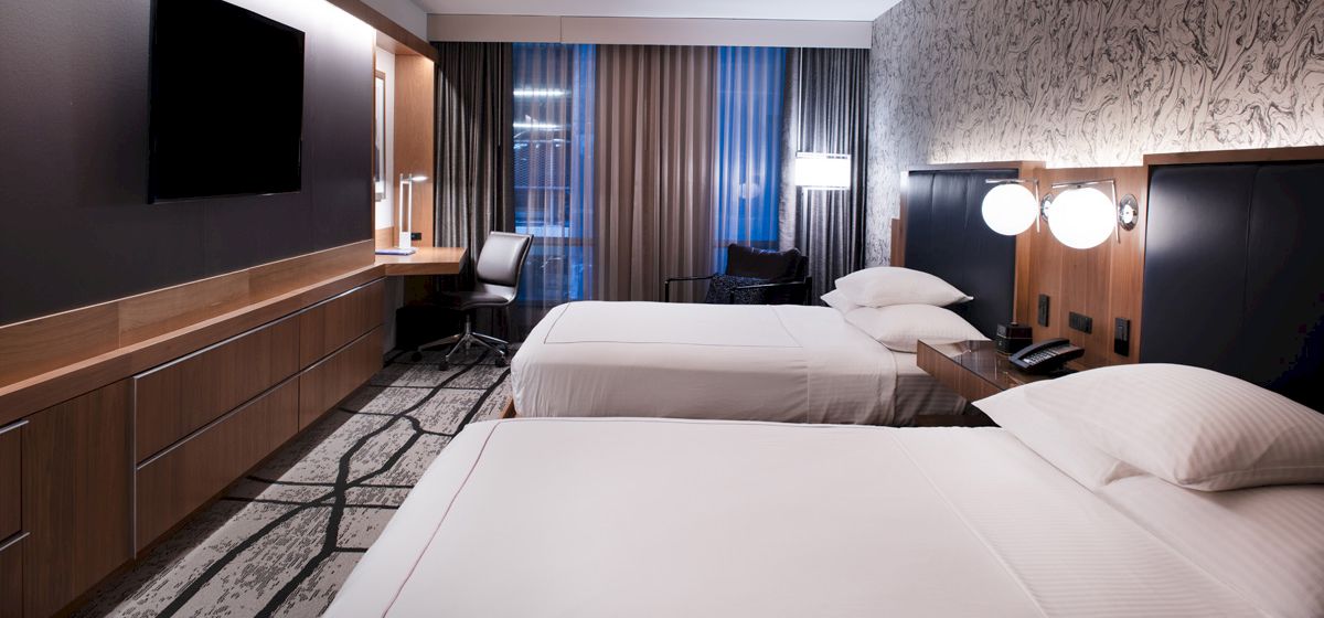A modern hotel room with twin beds, dark headboards, bedside lamps, and curtains beside a window.