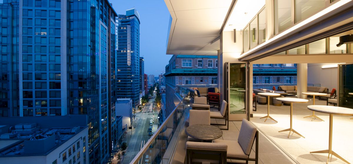 A modern high-rise terrace lounge with chairs overlooks a downtown city street between skyscrapers at dusk.